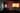 A photograph of a film projected in a dark space. The space is painted white with a dark ceiling and floor. The light in the room comes from a screen in the far corner, which is out of focus in the photograph. The screen shows an abstract yellow rectangular shape on an off which background, the edges appear to glow orange. Two vertical metal bars on the left of the image also reflect the light. Behing them, a speaker on a stand is visible.