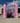 A photograph of an artwork presented outside in a paved urban space on a sunny day. The work consists of a large temporary structure, with two pink fabric wrapped columns on either side at the front. The word GAWP! is made out of large cut out letters fixed between the columns, and there is a fabric curtain under that is parted in the middle. Between the curtain, two women are standing with their arms round each other. They are smiling, and both wearing dark red tabards.