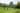 A group of 12 people are walking in a large circle in a lush green garden surrounded by trees. Some people are carrying large sticks. They are all wearing rain coats.