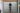 A person stood on a red step stool looks into a peep hole located on a shop shutter. The shutter has the words "LOOK HERE" and an arrow printed on it.
