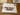 A photograph of a paper stencil with the words "what have we become" cut out. The stencil has been used, and there is masking tape around the edges, dark red paint around the letters, and the top right corder has been torn off.
