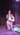 A photograph of a woman who is kneeing on a stage in front of a black curtain. She is wearing a black copped top and baggy dark coloured trousers and is holding a light-coloured cloth bag. She has brown skin and black hair, which is tied back, and her hands are in a prayer position. She is looking out beyond the camera.