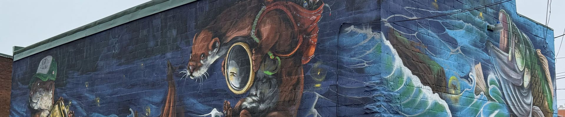 photo of the finished mural at M&J Supply. The mural features two otters playing in a river on one wall with a bass on the other wall. This mural was painted as part of the Active Arts Corridor Project.