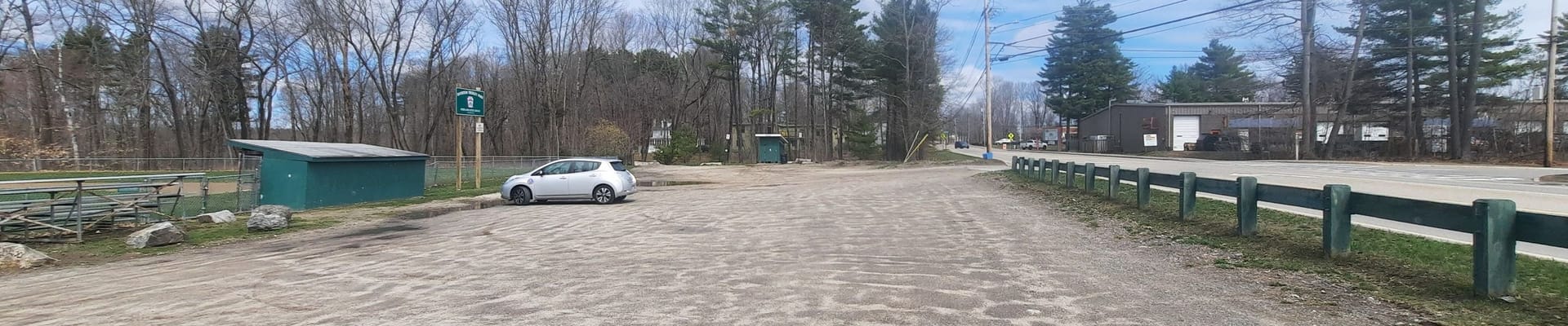 A dirt parking lot with fencing between the parking lot and road as well as a green dug out and baseball field on the left field. one single car parked in the parking lot.