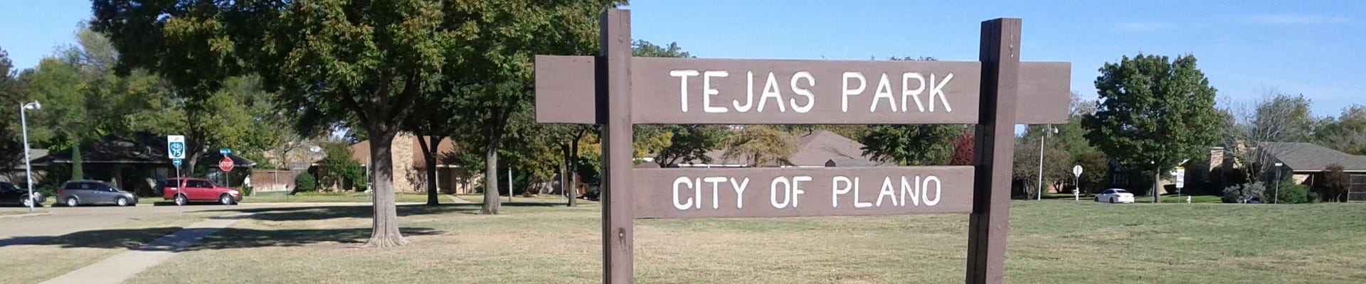 Brown sign displaying Tejas Park City of Plano on open grass field with trees near houses