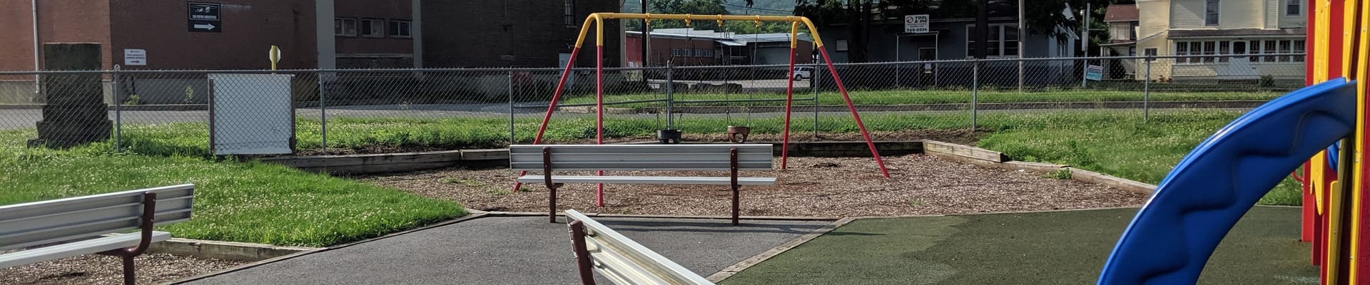 View of Piper-Harmon Playground in the City of Lock Haven. View includes a swing set, benches, and portion of a children-age jungle gym.