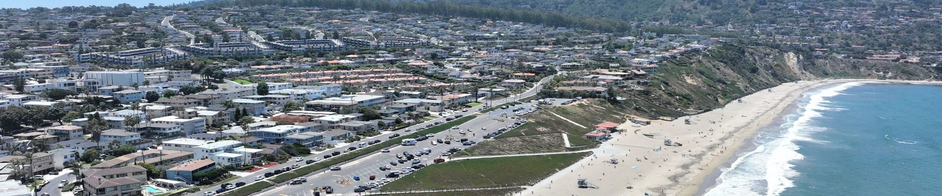 Aerial view of Torrance Beach showing the sandy shoreline, ocean waves, beachfront parking, and surrounding hillside residential neighborhood on a clear sunny day.