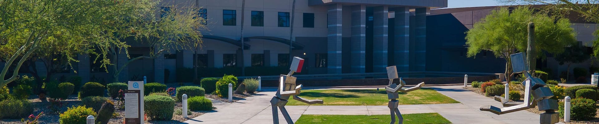 Modern civic building with landscaped courtyard featuring abstract metal sculptures of people reading books, set among desert plants, grass, and walkways under a clear blue sky.