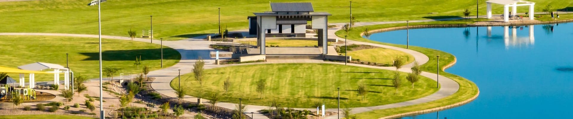 Aerial view of a large community park featuring shade structures, walking paths, grassy fields, and a blue lake with small ramadas and picnic areas.