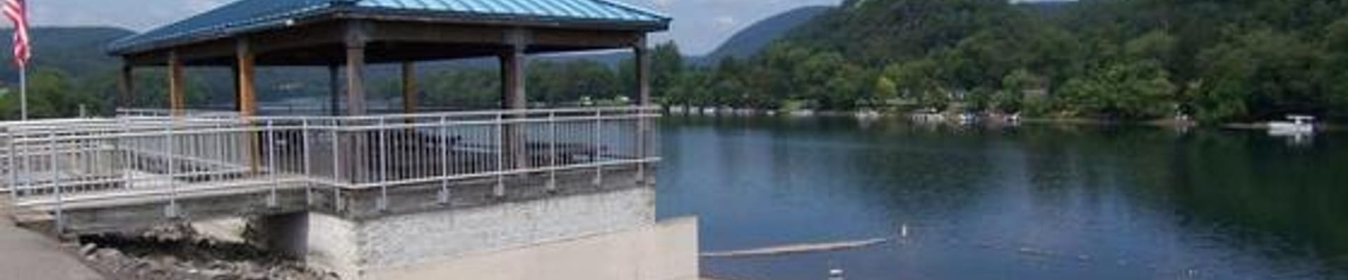 Photo of the Lock Haven City Beach. On the left is the levee and a pavilion with a blue roof, over-looking the steps and shore of the beach, and the Susquehanna River. There are mountains in the background with blue sky and light feathery clouds.