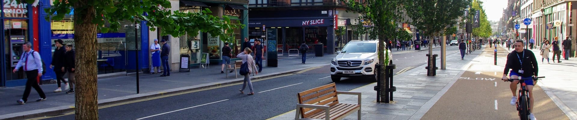 Image of a street with people walking, a cycle lane with someone cycling and a car on the road