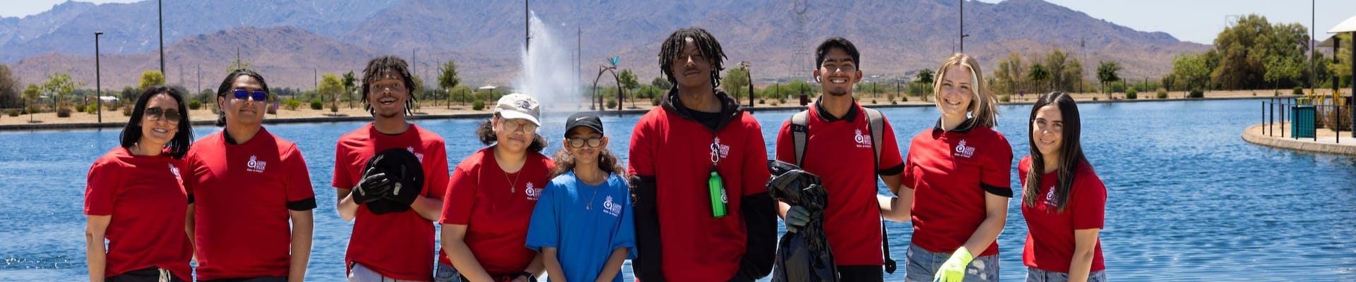 A group of volunteers wearing red shirts stands together by a lakeside park, holding trash bags and cleanup tools, with water and mountains visible in the background on a sunny day.