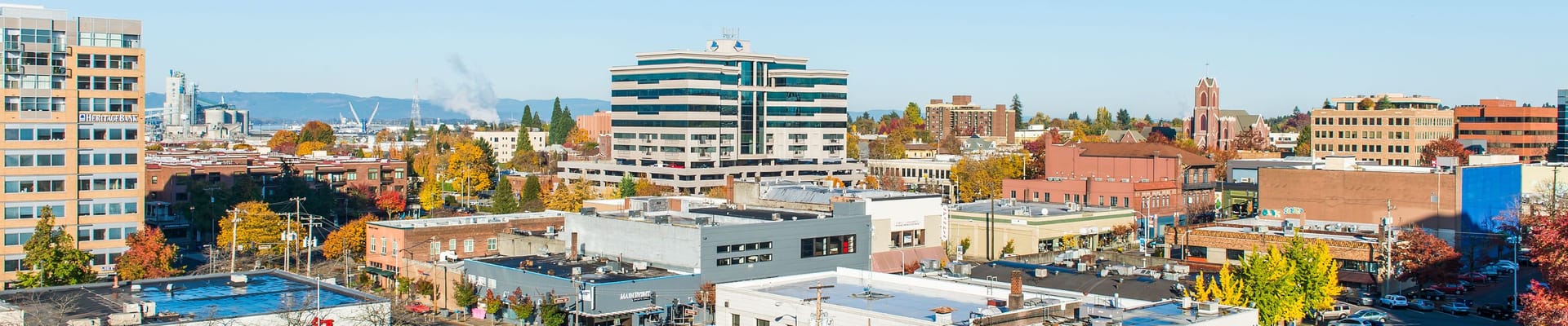 Panoramic view of 8th, Main, and Broadway downtown in autumn featuring tall buildings, low-rise rooftops, streets lined with orange and yellow trees, and industrial facilities visible in the distance beneath a cloudless blue sky.
