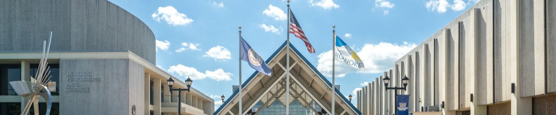 Wide exterior view of the Berglund Center in Roanoke, Virginia, with a covered central entrance, Roanoke flags, modern concrete buildings, and a bright blue sky.