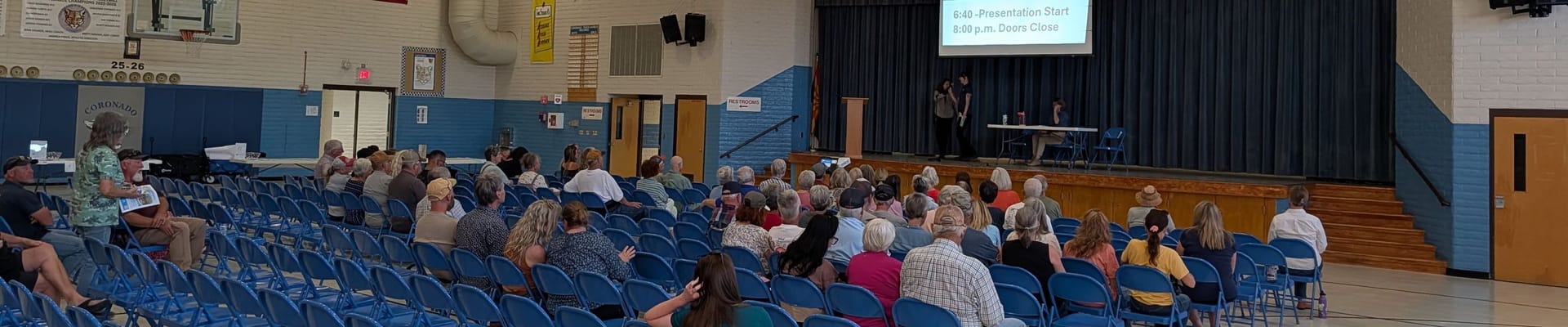 A gymnasium of blue chairs with people looking up at two presenters on stage.