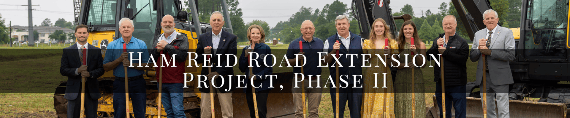 A group of officials stand holding shovels in a pile of sand in front of heavy machinery. A opaque black bar with white words reading, "Ham Reid Road Extension Project, Phase II" is shown.
