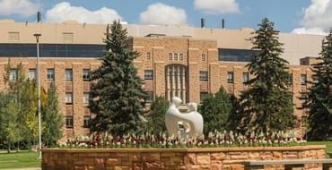 Landscaped plaza at the University of Wyoming with a white abstract sculpture surrounded by flowers, trees, and campus-style brick buildings in the background.