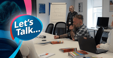 Three adult students sat at a collaborative large table taking notes whilst a female blonde haired coach is presenting information on a flipchart
