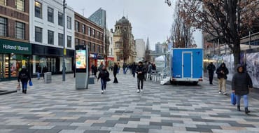 Photograph of a wide pedestrianised high street with high-quality paving, seating and street furniture. People are walking along the route, with shops and commercial buildings on either side. Digital information displays, mature trees and a historic landmark building are visible in the background, illustrating an attractive and accessible public realm designed to support town centre activity and movement.