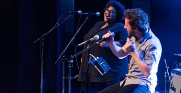 A man and woman sing into microphones on a stage. 