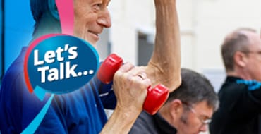 An older aged man smiling with dumbells in his hands pumping them upwards in the air.