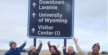 People posing beneath a directional wayfinding sign pointing toward Downtown Laramie, the University of Wyoming, and the Visitor Center.