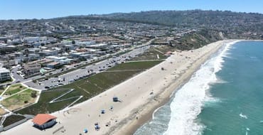 Aerial view of Torrance Beach showing the sandy shoreline, ocean waves, beachfront parking, and surrounding hillside residential neighborhood on a clear sunny day.