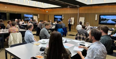 People sat around tables in a room at City Hall. They are facing a speaker at the front of the room. There are three screens showing the same image of a rive and buildings with the words 'London Local Nature Recovery Strategy' on.