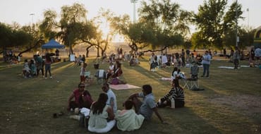 Families and park visitors gather on the grass at Eldorado Park during an outdoor community event, with trees and the setting sun in the background.