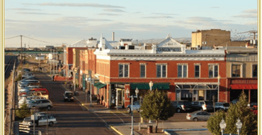 View of historic downtown Laramie with brick storefronts, parked cars, street trees, and decorative lighting along a commercial street.