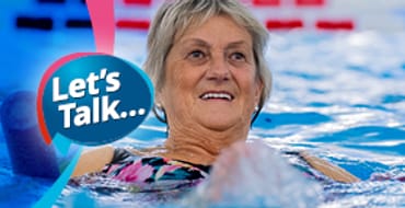 An older aged grey haired lady leaning back on a blue noodle float in a swimming pool with a smile on her face.