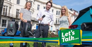 A photo of three people smiling and plugging an electric car charger into a car with Let's Talk branding