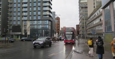 Street-level view of Ilford Hill showing modern residential development alongside a busy road with buses, vehicles and pedestrians. The image illustrates higher-density development, public transport accessibility and an urban arrival route into the town centre.