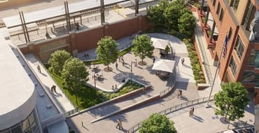 Aerial view of a landscaped public plaza next to a brick building and train station, with trees, seating areas, walkways, ramps and people walking and biking.