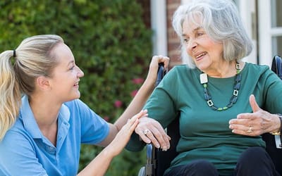 A middle aged lady with blonde hair crouched down looking at an older aged lady in a wheelchair and both are smiling and understanding each other.