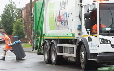 A rubbish collection wagon is in the middle of the shot with a man in orange highly visible clothing in front. He is pulling a blue topped wheelie bin back to where he collected it.