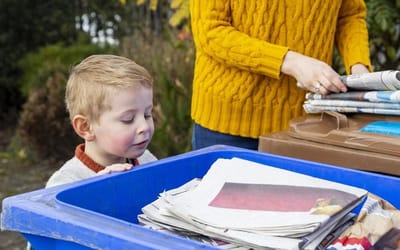 A young boy looking down into a blue outdoor rubbish wheelie bin with a responsible adult beside him with a hand full of paper materials.