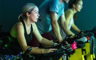 A lady, a man and a woman in a row riding indoor bicycles participating in a fitness class.