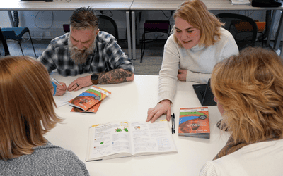 Four students on a table around a book helping each other learn a new skill