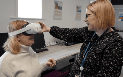 Two blonde haired women, one a student and one a teacher, in a modern classroom surrounded by computers. The teacher is placing a VR headset over the eyes of the student.