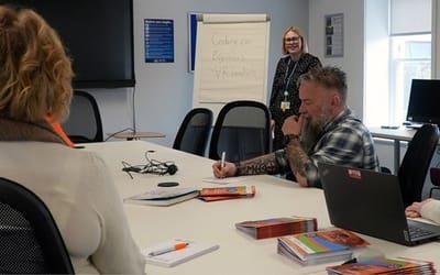 A teacher with blonde hair is stood in front of a flip chart that reads 'Coding for beginners...' There are four students spread around a business meeting table listening and taking notes.