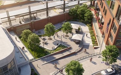 Aerial view of a landscaped public plaza next to a brick building and train station, with trees, seating areas, walkways, ramps and people walking and biking.