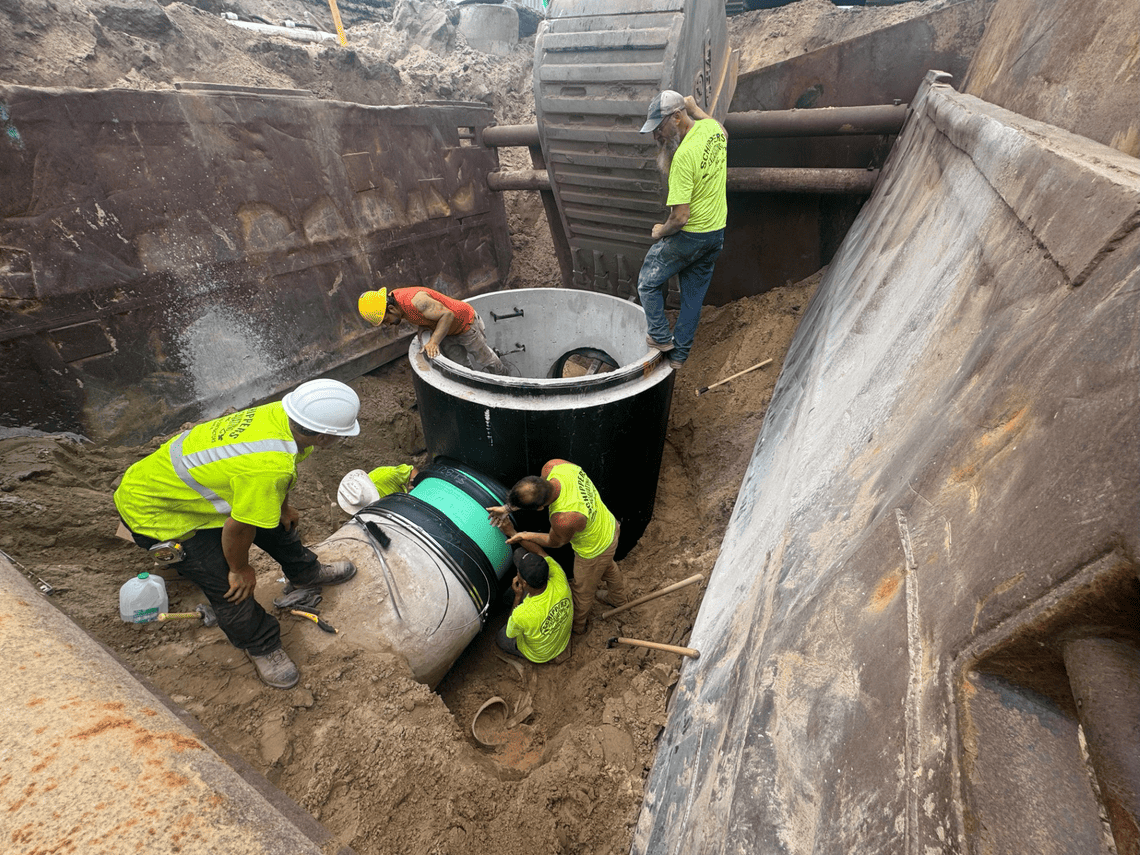 Construction workers install a new sanitary sewer manhole and underground pipe inside a reinforced excavation.