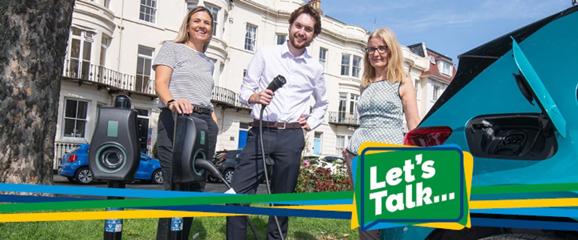 A photo of three people smiling and plugging an electric car charger into a car with Let's Talk branding