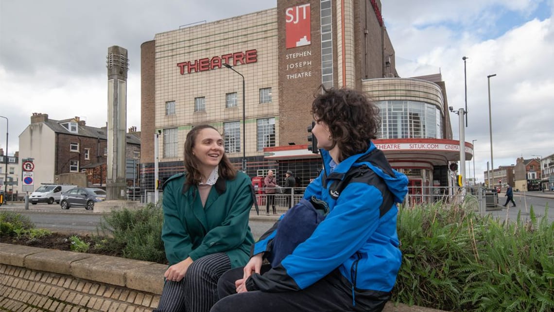 Two young women sat on the wall outside Scarborough train station with the Stephen Joseph Theatre behind them facing each other talking.