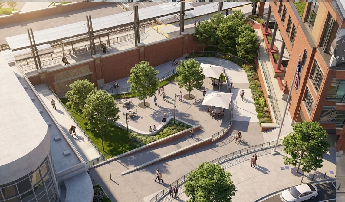 Aerial view of a landscaped public plaza next to a brick building and train station, with trees, seating areas, walkways, ramps and people walking and biking.