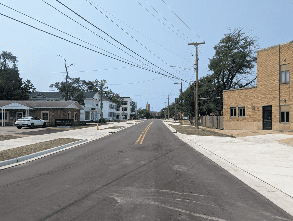 Newly paved roadway with new concrete sidewalks, curbs, and driveways along both sides of the street. Utility poles and overhead wires line the road, with a mix of residential and commercial buildings visible in the distance.