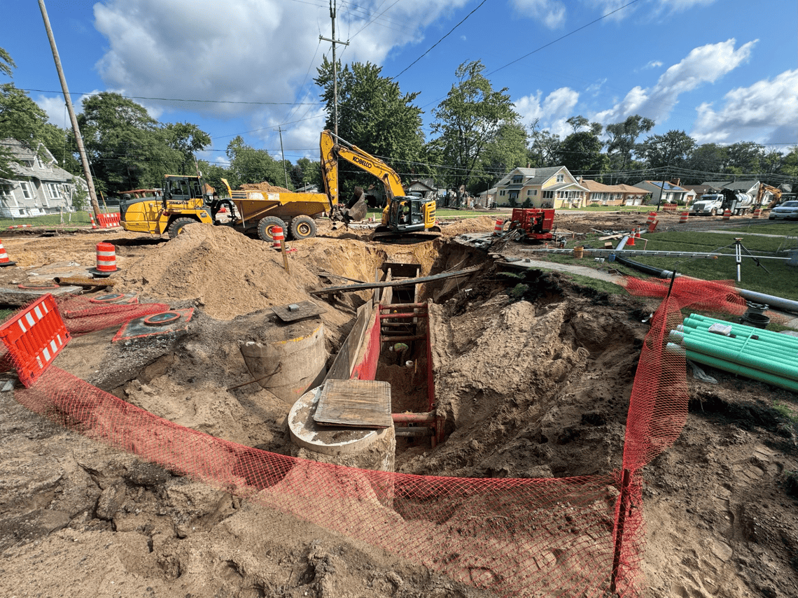 Construction crew works in a deep trench installing new sanitary sewer infrastructure on Oak Grove, with excavators, utility pipes and safety barriers surrounding the work area.