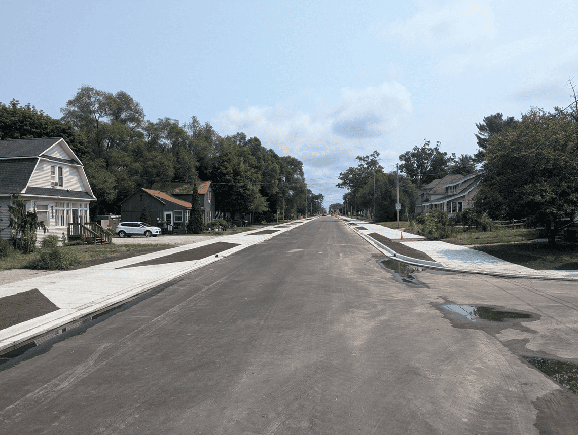 Newly paved residential street with new concrete sidewalks, curbs, and driveway approaches. Homes, mature trees, and utility lines line both sides of the roadway, with construction activity visible in the distance.