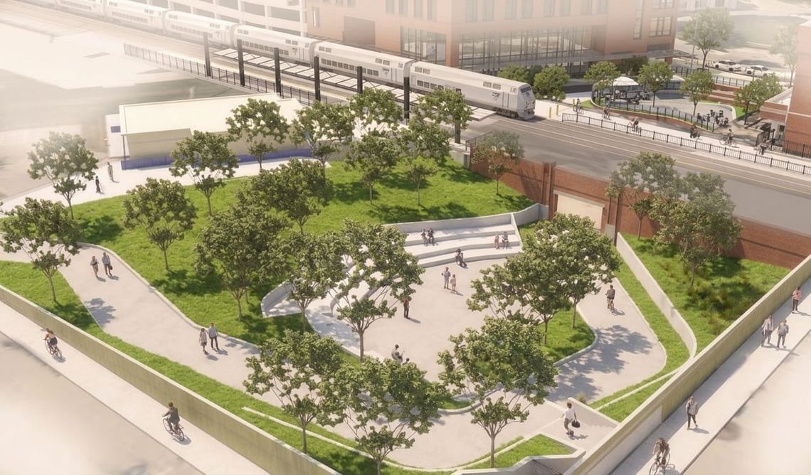 Aerial view of a landscaped public plaza with walking and biking paths, mature trees, grassy areas and tiered seating. A train is visible on the tracks beyond the plaza, with a street and buildings in the background.